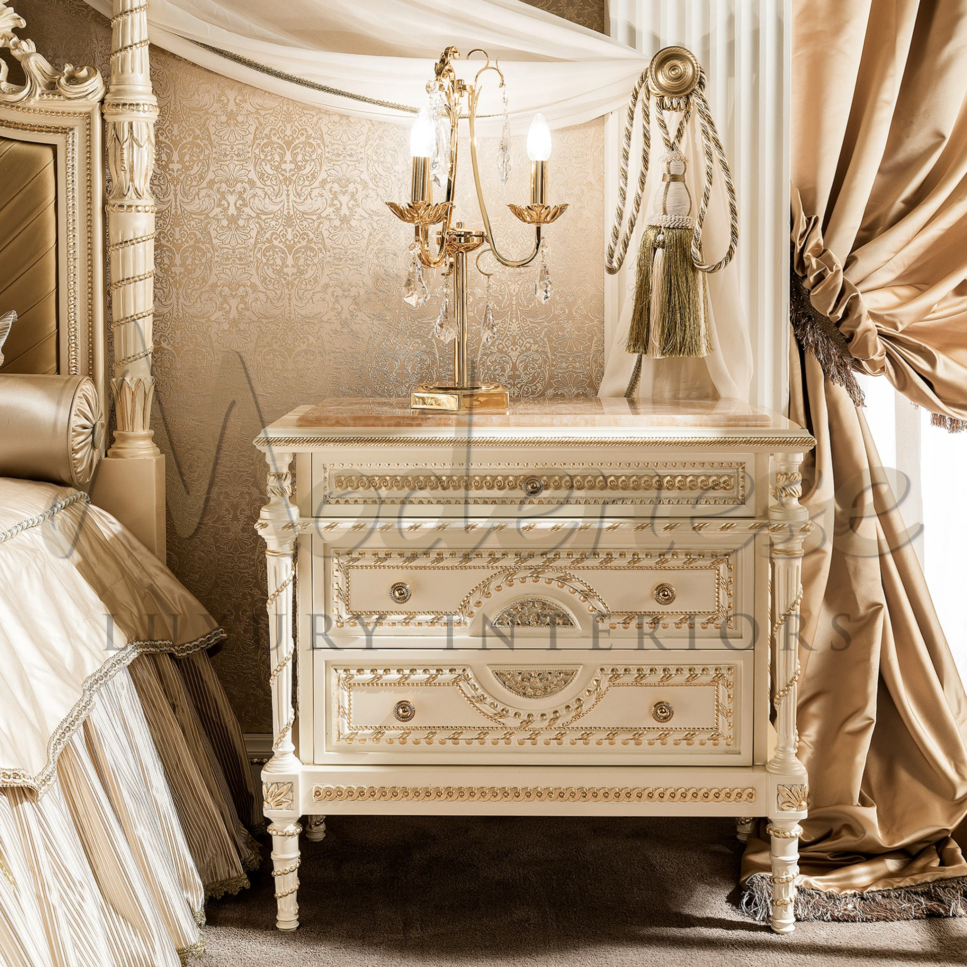 A luxurious cream-colored nightstand by Modenese Furniture, adorned with intricate gold detailing and a marble surface, placed alongside elegant drapery and a classic candelabra-style lamp.