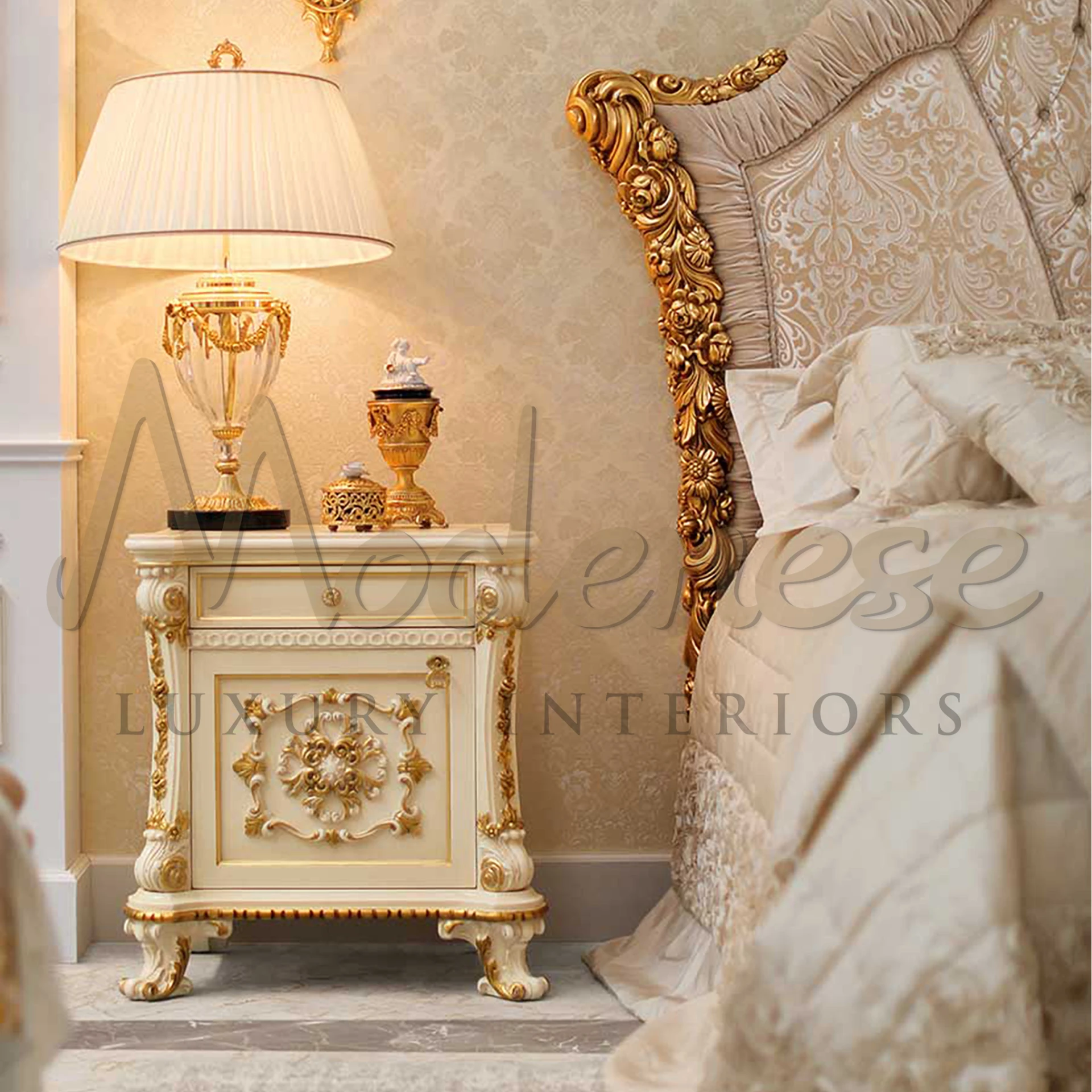 A baroque-style cream nightstand with gold trim, topped with a lamp and ornate decorative items, beside an opulent bed.Made by Modenese Furniture Manufacturer.