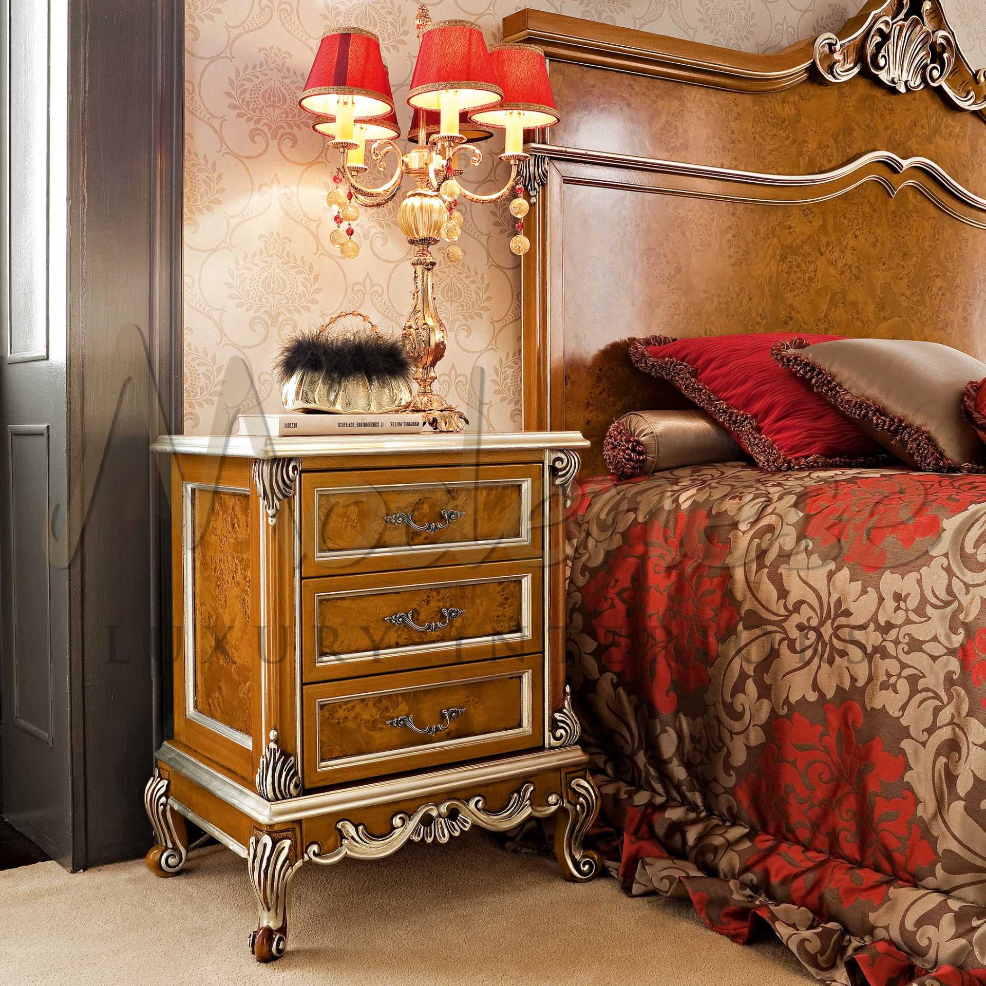 Traditional nightstand with burl wood finish and ornate metal details, beside a bed with rich red and beige patterned bedding, under a red-shaded candelabra lamp.