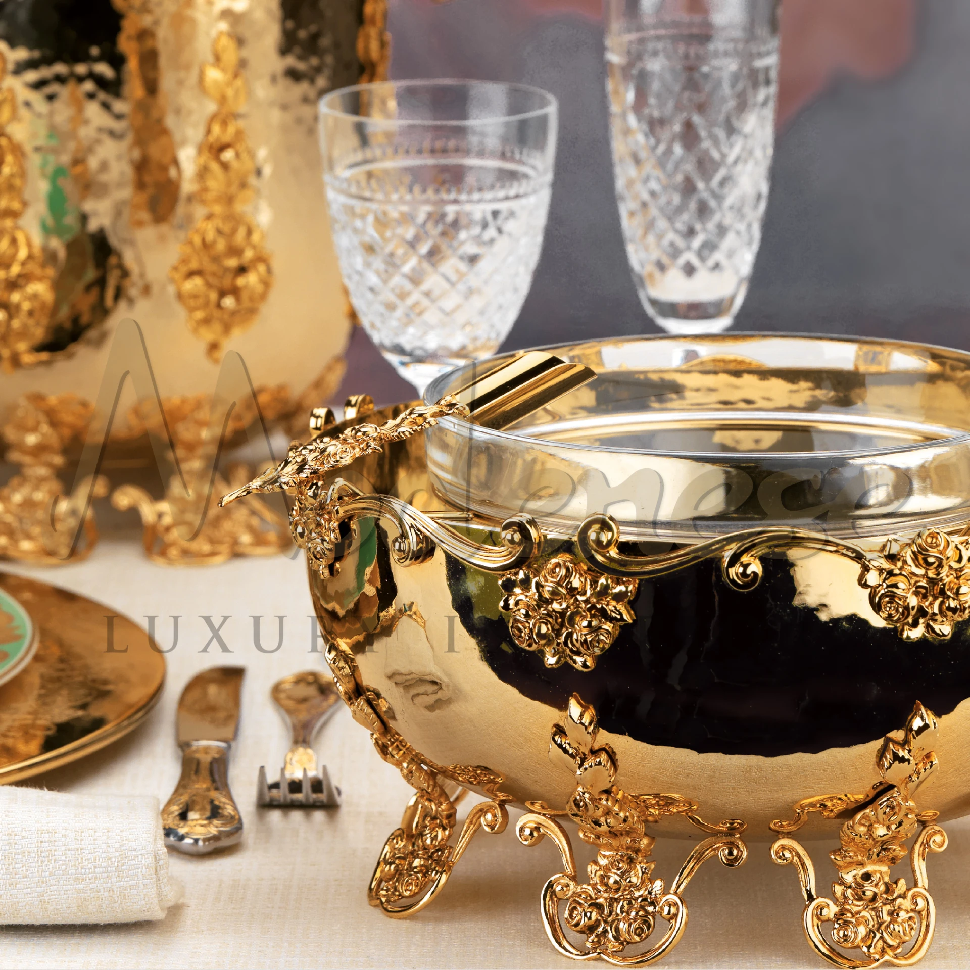 A gold Caviar holder with spoon on a table next to glasses and utensils.